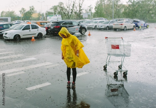 Girl in yellow raincoat stand alone under rain in parking lot. Look down on wet asphalt. Many cars behind. Empty grossery cart beside. Rainy weather. photo