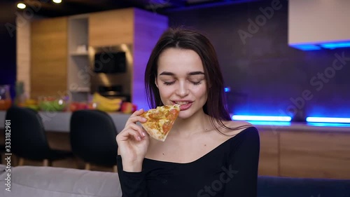 Good-natured smiling young brunette sitting on the couch in the contemporary room and eating pizza,posing on camera photo