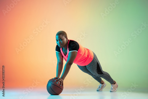 Young african-american plus size female model's training on gradient background in neon light. Doing workout exercises with fit ball. Concept of sport, healthy lifestyle, body positive, equality. photo