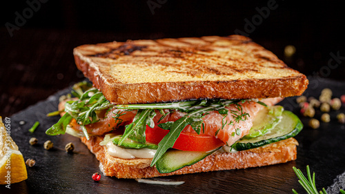 Vegetarian American Cuisine. Homemade toast sandwich with red fish salmon, arugula, cucumbers and tomatoes, white sauce. Sandwich on a black background. copy space text