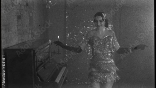 Young happy beautiful woman in bright sparkling luxury silver dress, smiles gown fluttering dances in room near piano, black and white old fashioned film with added noise. 20s stylized carnival party photo