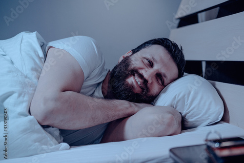 A lonely bearded dark-haired man with a very happy face lies on his side and looks at the phone at night in bed. Dreams, a date succeeded, happy, white bedding.
