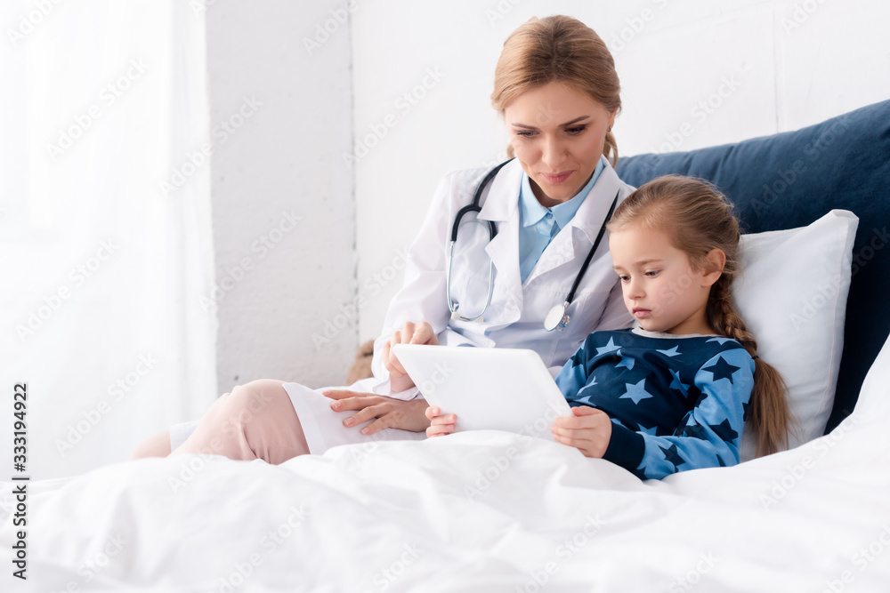 attractive doctor in white coat sitting near sick kid with digital tablet attractive doctor in white coat sitting near sick kid with digital tablet