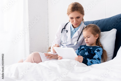 attractive doctor in white coat sitting near sick kid with digital tablet