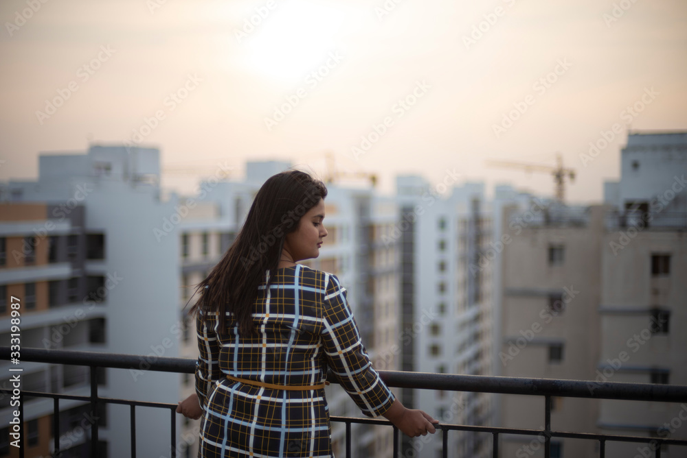 Back side of an young brunette Indian Bengali brunette plus size woman in western dress standing on rooftop in urban background while her hair is blowing in wind during sunset. lifestyle and fashion.