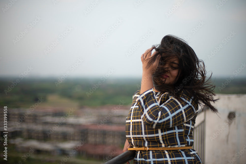 An young brunette Indian Bengali brunette plus size woman in western dress standing on rooftop/balcony in urban background while her hair is blowing in wind during sunset. lifestyle and fashion.