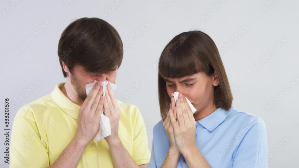 young couple in yellow and blue shirt blow noses into tissues on grey background