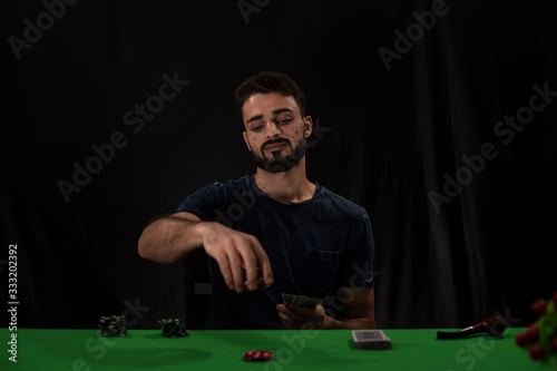 Portrait of young brunette Indian Kashmiri man in casual tee shirt playing cards on a casino poker table in black copy space studio background. lifestyle and fashion.