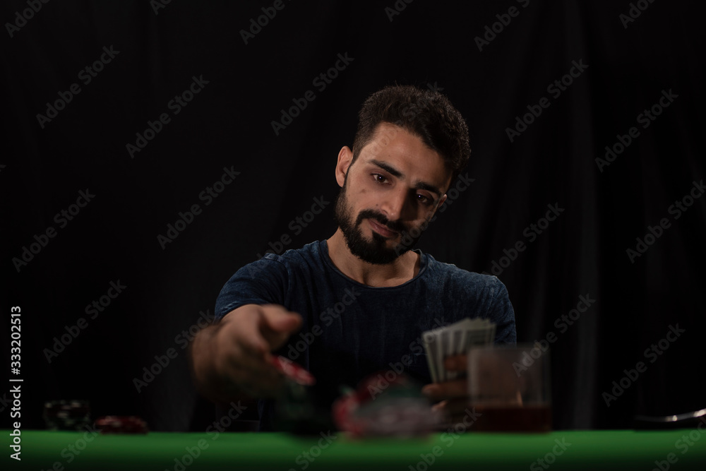 Portrait of young brunette Indian Kashmiri man in casual tee shirt playing cards on a casino poker table in black copy space studio background. lifestyle and fashion. Portrait of young brunette Indian Kashmiri man in casual tee shirt playing cards on a casino poker table in black copy space studio background. lifestyle and fashion.