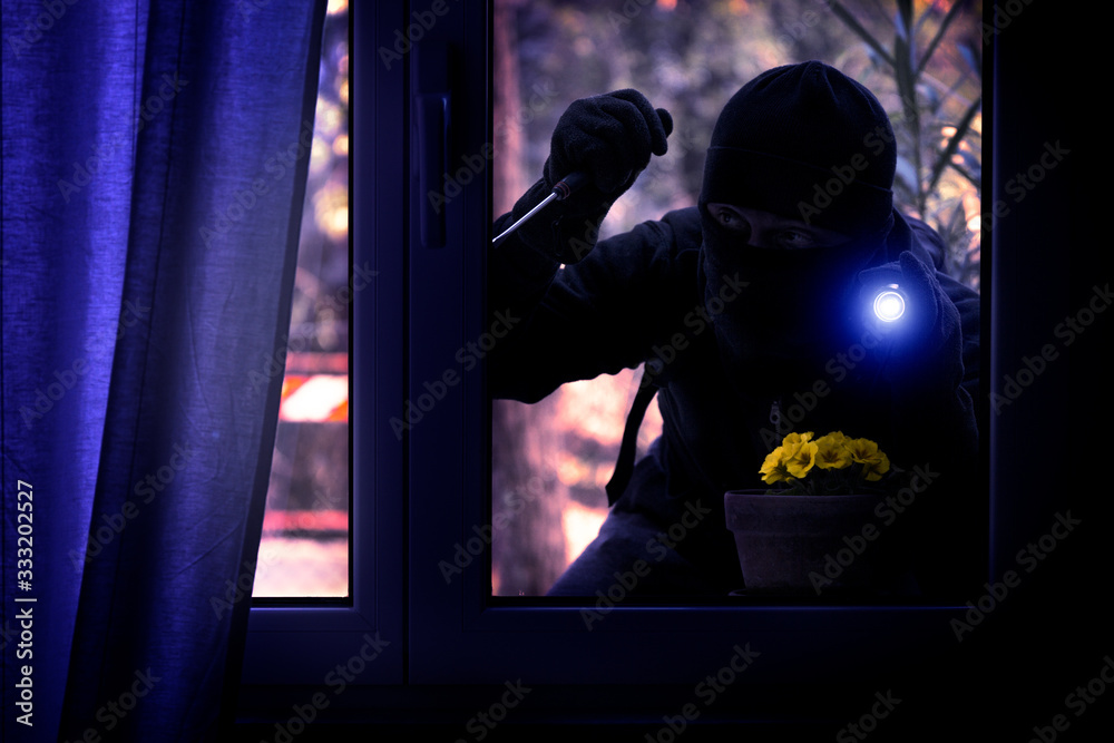 Hooded burglar forcing the window frame - Silhouette of thief with screwdriver and flashlight is breaking into the apartment - Concept of intrusion and danger view from indoor dark shaded image