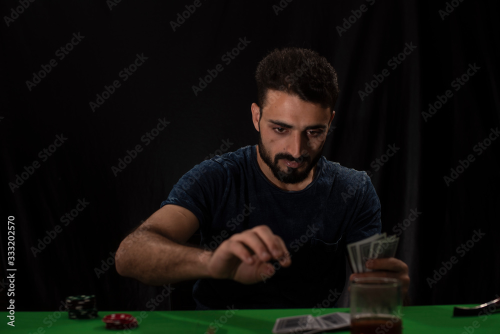 Portrait of young brunette Indian Kashmiri man in casual tee shirt playing cards on a casino poker table in black copy space studio background. lifestyle and fashion. Portrait of young brunette Indian Kashmiri man in casual tee shirt playing cards on a casino poker table in black copy space studio background. lifestyle and fashion.