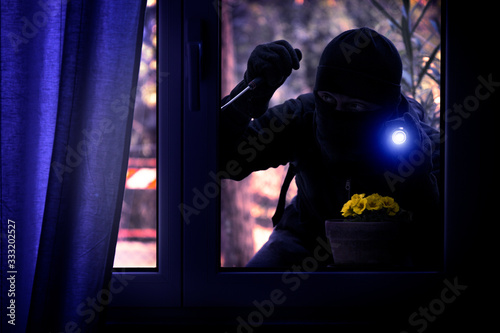 Hooded burglar forcing the window frame - Silhouette of thief with screwdriver and flashlight is breaking into the apartment - Concept of intrusion and danger view from indoor dark shaded image