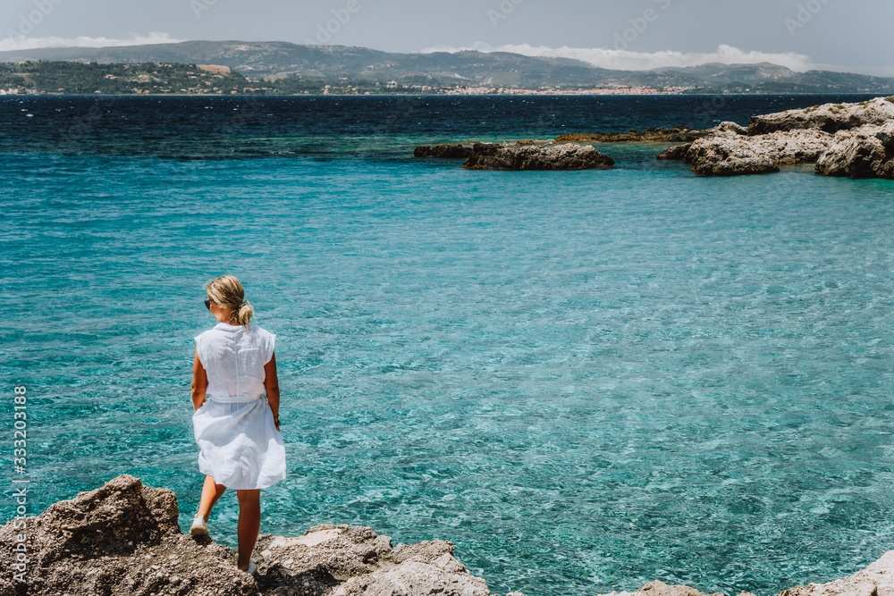 Adult female in white dress on summer vacation enjoying sea coast landscape of small beach with crystal clear blue water. Greece, Kefalonia