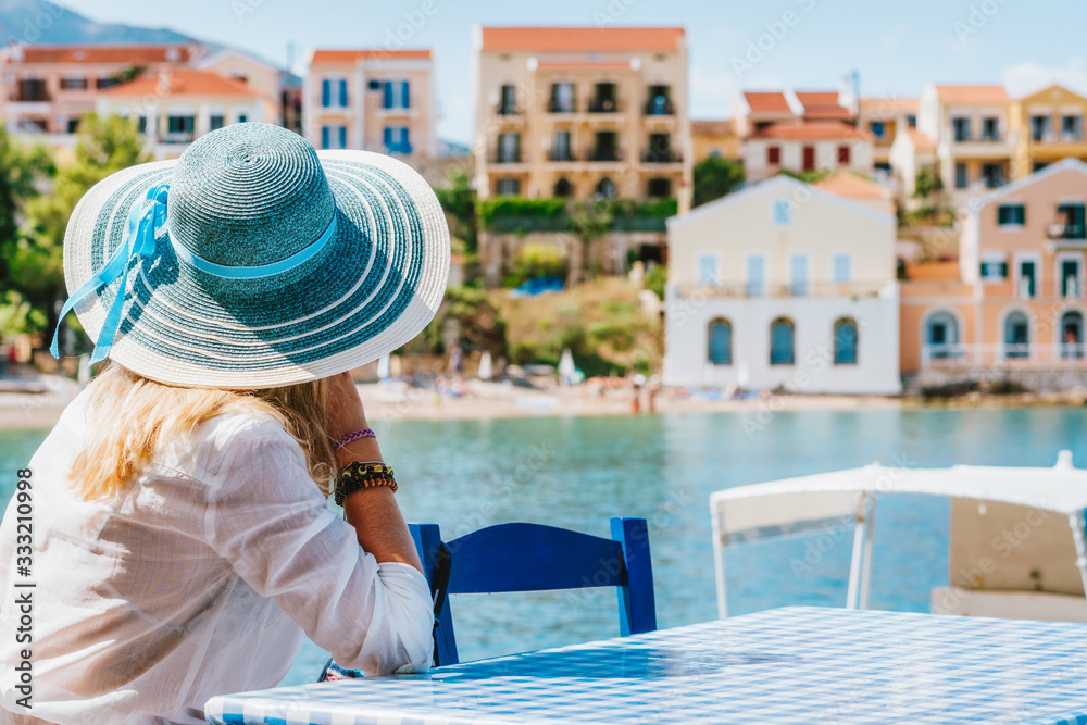 Rear view of tourist woman sitting cafe wear blue sunhat admire colorful tranquil houses of Assos village on sunny day. Visiting Kefalonia in summer. Travel vacation in Greece