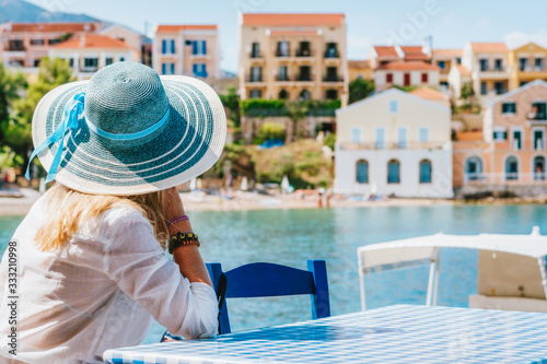 Rear view of tourist woman sitting cafe wear blue sunhat admire colorful tranquil houses of Assos village on sunny day. Visiting Kefalonia in summer. Travel vacation in Greece