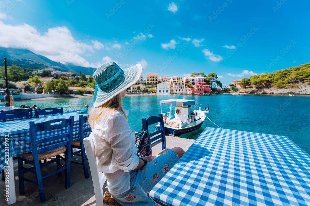 Naklejka premium Holiday vacation. Woman in cafe enjoying time in Assos village in front of emerald bay of Mediterranean sea white boat and beautiful traditional houses of in Kefalonia, Greece