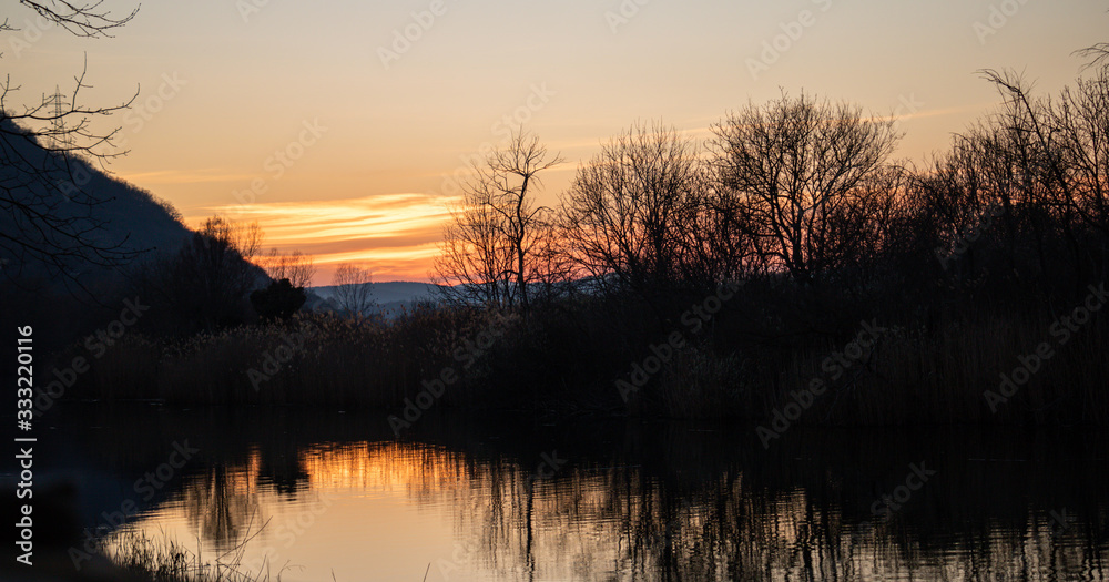 Sunrise, sunset on lake. Silhouette of trees reflected on the pond and colorful sunbeams colorize the water. Sky and mountains backdrop. Wallpaper.