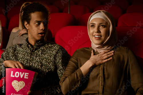 African american muslim man with his wife sitting in movie theater, watching movie, eating popcorn, smiling. © Infot