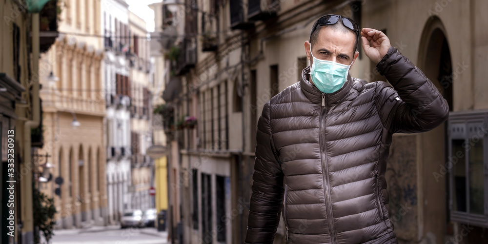 uomo scuro con giubotto e occhiali da sole in testa, indossa una mascherina protettiva chirurgica mentre sta in giro per la città con il trolley