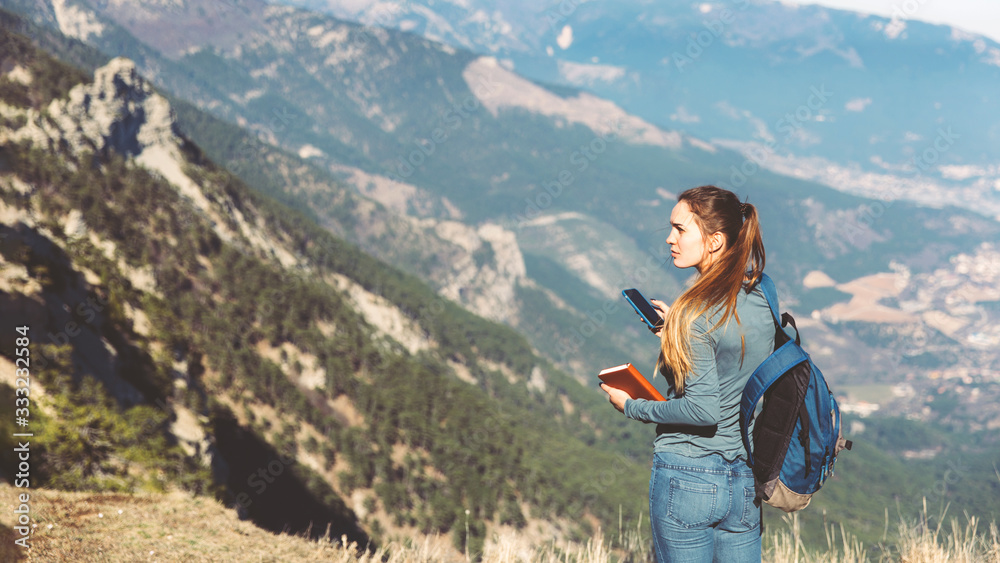 Naklejka premium Tourist girl making a photo shoot of mountain. Young beautiful girl travels alone in the mountains in spring or autumn, looks into the distance and enjoys nature, rocks and green forests, view of the
