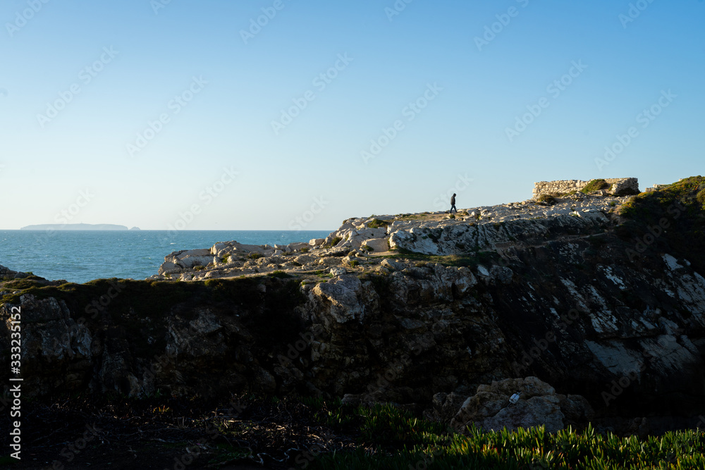 Man walking on top of a rocky mountain on the coastline with the ocean in the background.