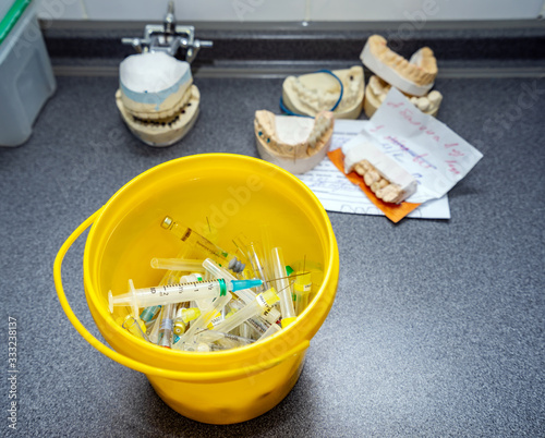 Medical waste, syringes with needles and empty vials in the trash bucket closeup. Dental forms and models with prosthetic teeth in the background photo