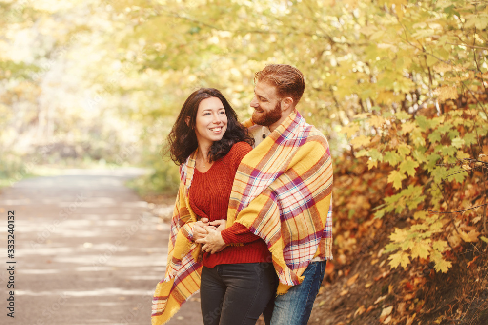 Fototapeta premium Beautiful couple man woman in love. Boyfriend and girlfriend wrapped in yellow blanket plaid hugging in a park on autumn fall day. Tenderness and happiness. Authentic real people.
