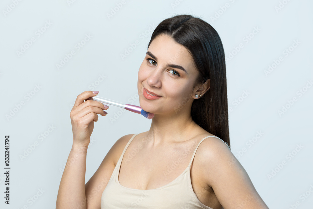 Close-up of a young girl in a light top on a white background making a facial make-up. A pretty woman holds a cosmetic brush near her face and smiles. Close-up of a young girl in a light top on a white background making a facial make-up. A pretty woman holds a cosmetic brush near her face and smiles.