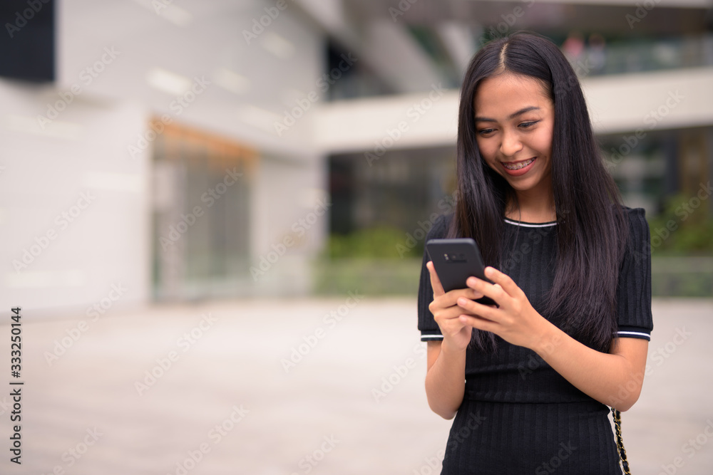 Happy young beautiful Asian tourist woman using phone at the mall outdoors