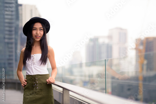 Happy young beautiful Asian tourist woman thinking against view of the city