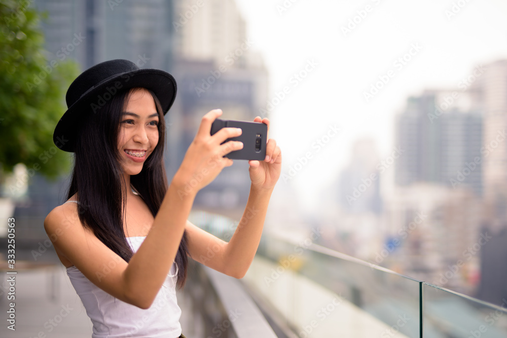 Happy young beautiful Asian tourist woman taking picture with phone against view of the city