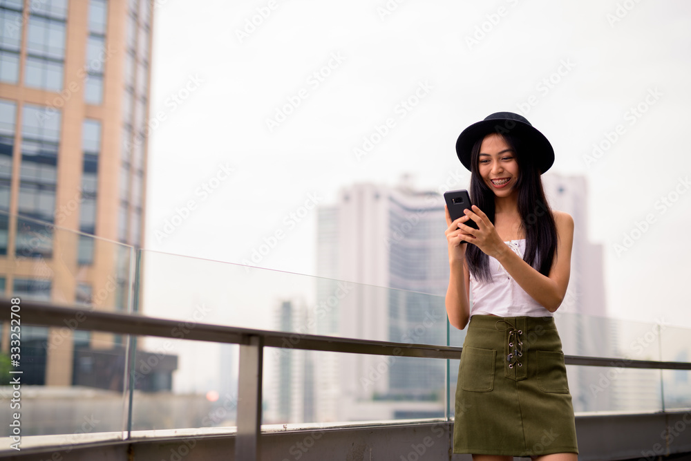 Happy young beautiful Asian tourist woman using phone against view of the city
