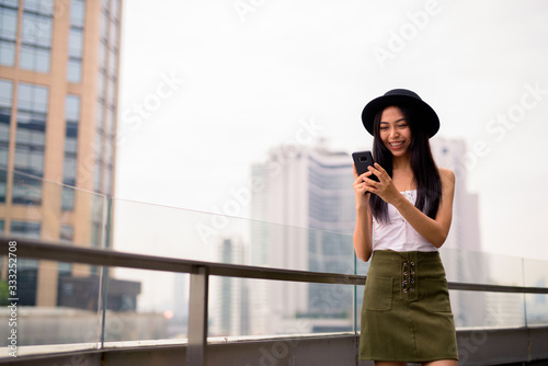 Happy young beautiful Asian tourist woman using phone against view of the city