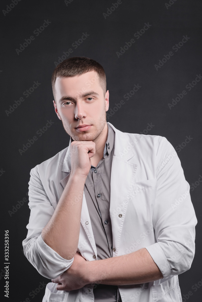 Young, pretty man in a medical coat on a gray background. The concept of medical education and medical activity Young, pretty man in a medical coat on a gray background. The concept of medical education and medical activity
