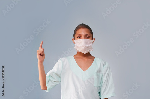 A young nurse or assistant in medical clothing and a mask looks into the frame.