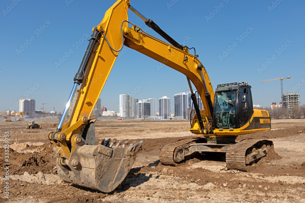 Heavy excavator at construction site with working industrial equipment against urban scene Heavy excavator at construction site with working industrial equipment against urban scene
