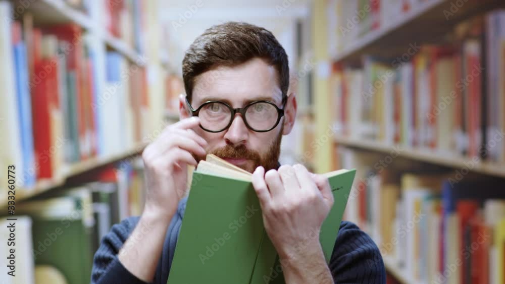 Playful handsome young man reading a book looking around a library room sitting between bookshelves. Real portrait of flirty bearded librarian on background. Playful handsome young man reading a book looking around a library room sitting between bookshelves. Real portrait of flirty bearded librarian on background.