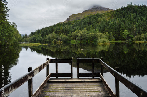 Fototapeta Naklejka Na Ścianę i Meble -  Landscape, reflections and a wooden walkway upon Glencoe Lochan on a cloudy day, in the Scottish Highlands, Scotland. 