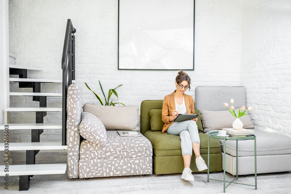 Young creative and well-dressed woman working on a digital tablet while sitting on the comfortable couch at home. Interior view on the modern and bright living room Young creative and well-dressed woman working on a digital tablet while sitting on the comfortable couch at home. Interior view on the modern and bright living room