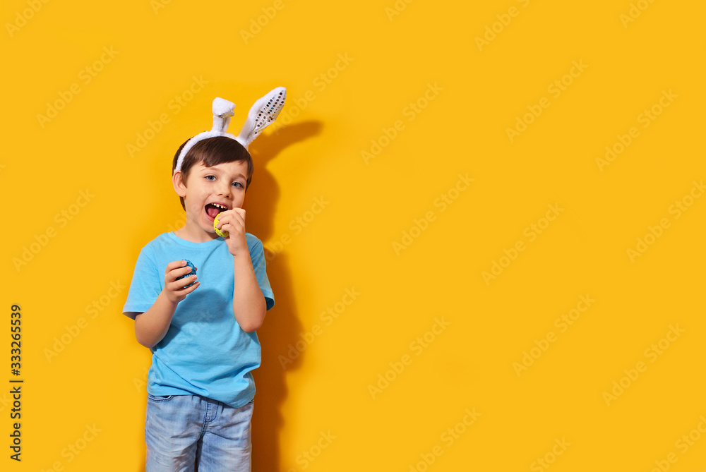 Naklejka premium Portrait of a smiling adorable little boy with bunny ears and holding colorful easter eggs isolated orange background