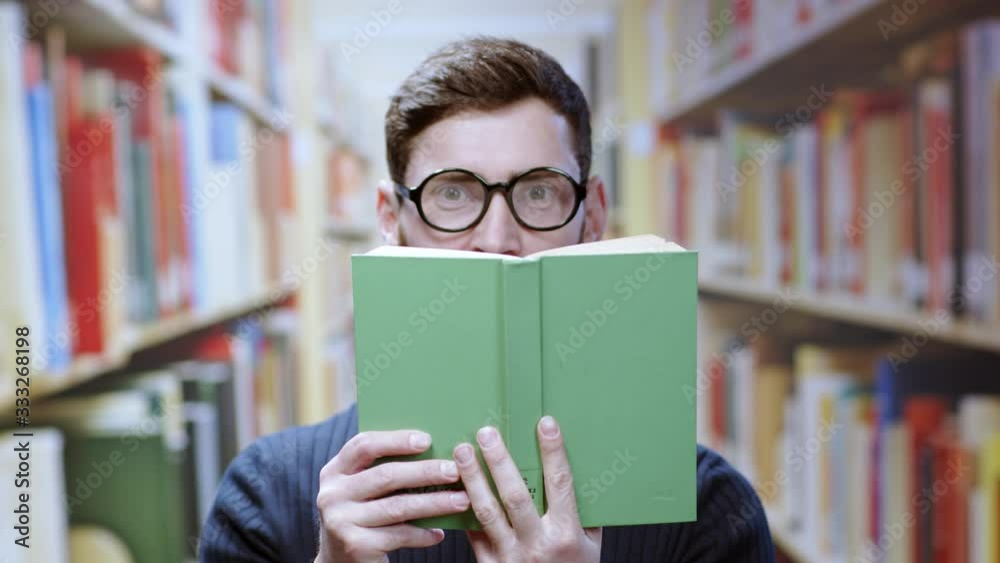 Portrait of shy clever young bearded man staying in the library hiding behind the book spying look at camera scared. Comedy, fun, entertainment. Portrait of shy clever young bearded man staying in the library hiding behind the book spying look at camera scared. Comedy, fun, entertainment.