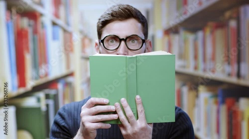 Shy unsociable bearded smart young man student wearing glasses hiding behind a green book scared of people staying in college library. photo