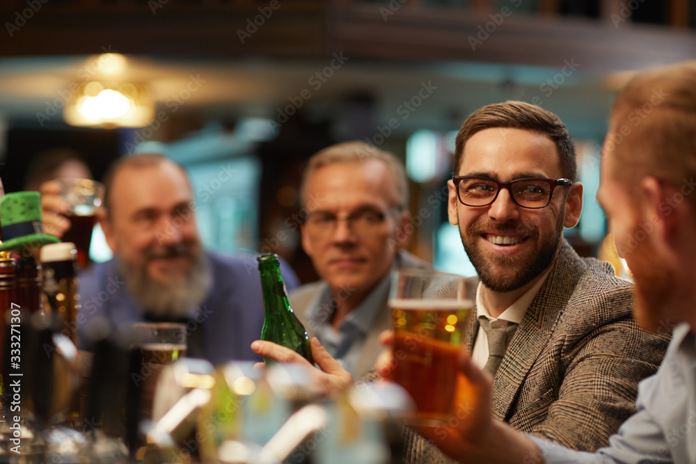 Bearded young man in eyeglasses smiling and talking to his friends while they sitting in the bar and drinking beer