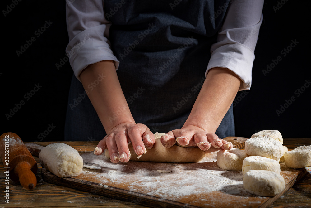 Female hands knead the dough on a wooden antique table on a dark background, close-up, shallow depth of field, beautiful directional lighting. Concept of home baking and comfort.