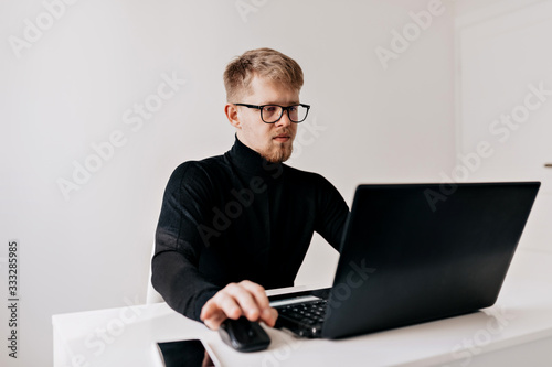 Young man team leader. Confident young man working at his desktop with laptop and looking at camera with smile in his light office