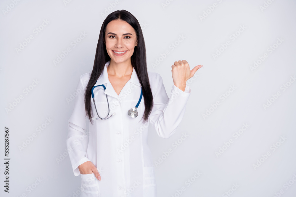 Photo of beautiful doctor lady good mood smiling patients direct thumb finger empty space insurance offer wear white lab coat stethoscope isolated grey color background Photo of beautiful doctor lady good mood smiling patients direct thumb finger empty space insurance offer wear white lab coat stethoscope isolated grey color background