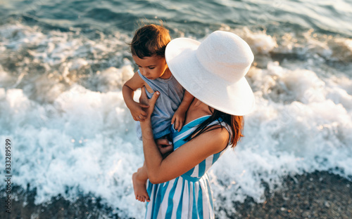 Fototapeta Naklejka Na Ścianę i Meble -  Horizontal top view image of pretty young mother playing with her daughter at the seaside, wearing blue striped dress and white hat background. Female playing with cheerful little girl at ocean sunset
