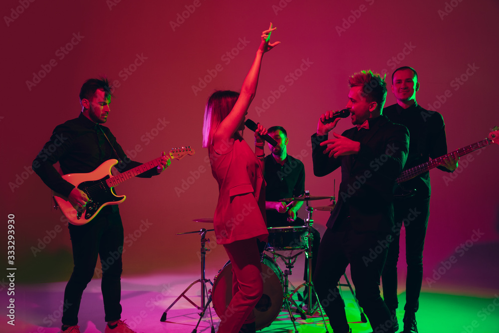 Young and joyful caucasian musicians performing on red studio background in neon light. Concept of music, hobby, festival. Colorful portrait of modern artist. Attented and inspired. Art, cover band.