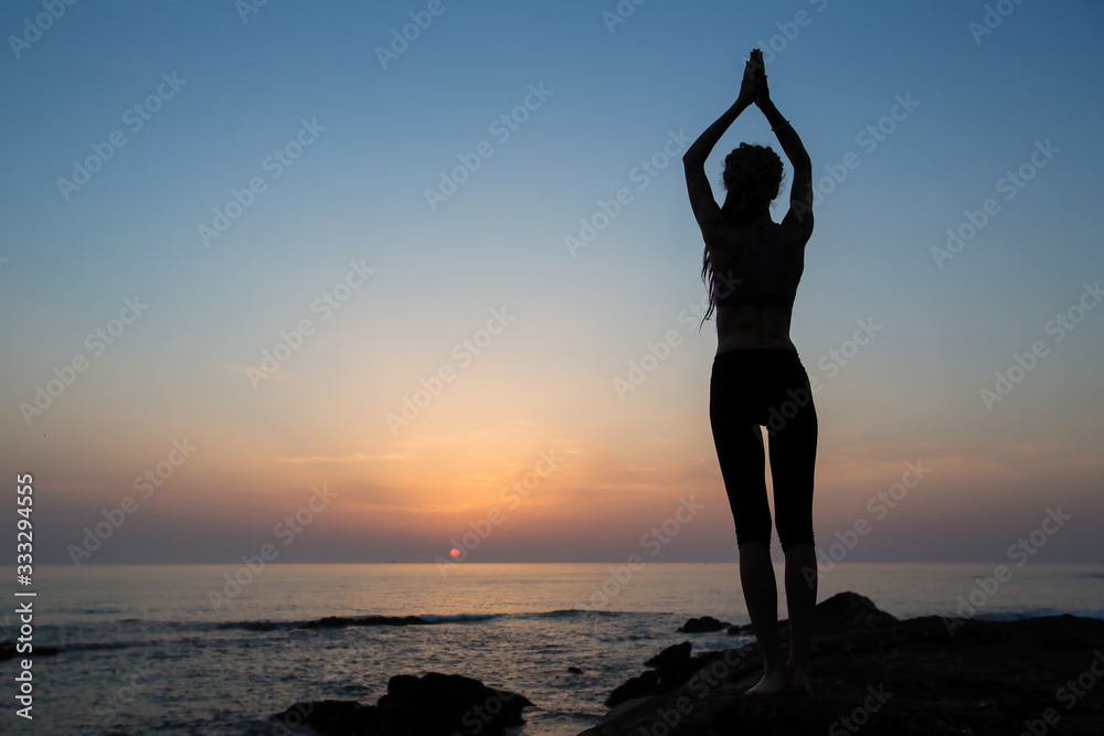 Silhouette of a flexible yoga woman stands on the ocean shore at evening.