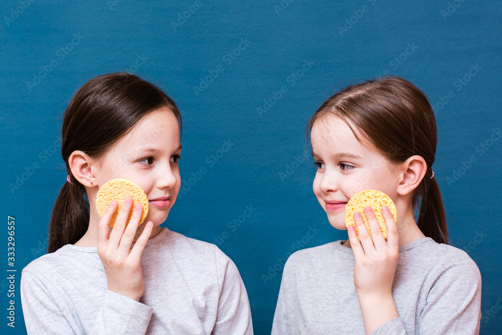 Two girls wash themselves with face sponges, smile and look at each other on a blue background. Child Hygiene Concept Two girls wash themselves with face sponges, smile and look at each other on a blue background. Child Hygiene Concept